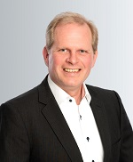 Ein Projekt Manager mit kurzem, blondem Haar, einem weißen Hemd und einem schwarzen Sakko steht vor einem hellen Hintergrund.
