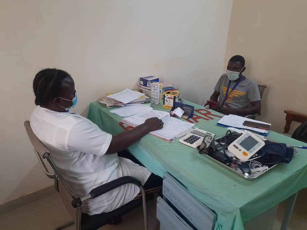 Zu sehen ist ein Arzt und ein Patient aus Burkina Faso, die sich gegenüber an einem Tisch mit unterschiedlichen Untersuchungsgeräten und Unterlagen sitzen.