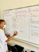 The picture shows a man standing at a whiteboard, writing and explaining something.