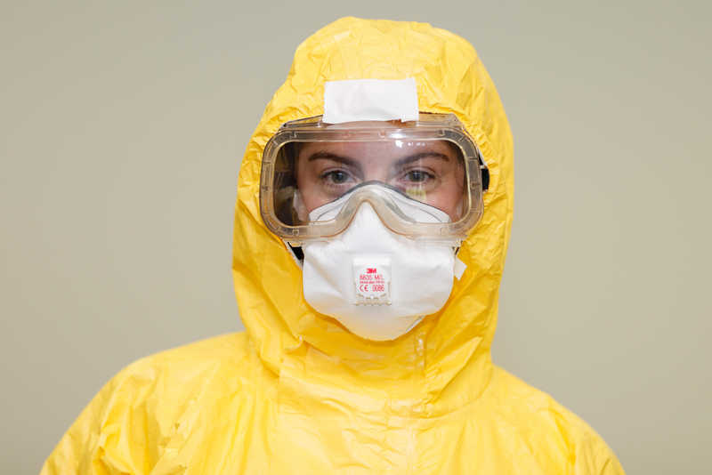 A friendly smiling European woman in a yellow biosafety suit poses for a single photo. She is wearing a protective mask and goggles.