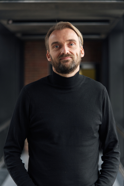 PD Dr. Thomas Jacobs: ein Forscher mit kurzem, dunklen Bart, blonden längeren Haaren in schwarzer Kleidung vor dunklem Hintergrund.